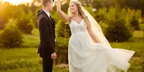 A bride and groom dancing in a grassy field at sunset, with the bride's dress and veil flowing in the wind.