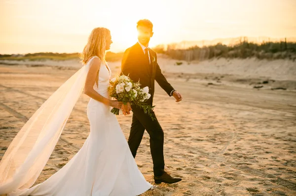 A bride and groom walking hand-in-hand on a sandy beach during golden hour, with the bride holding a bouquet.