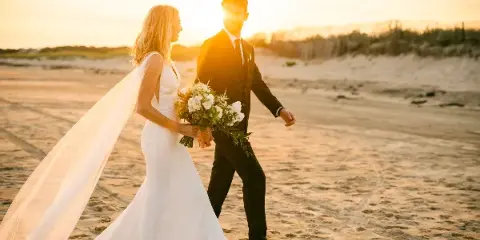 A bride and groom walking hand-in-hand on a sandy beach during golden hour, with the bride holding a bouquet.