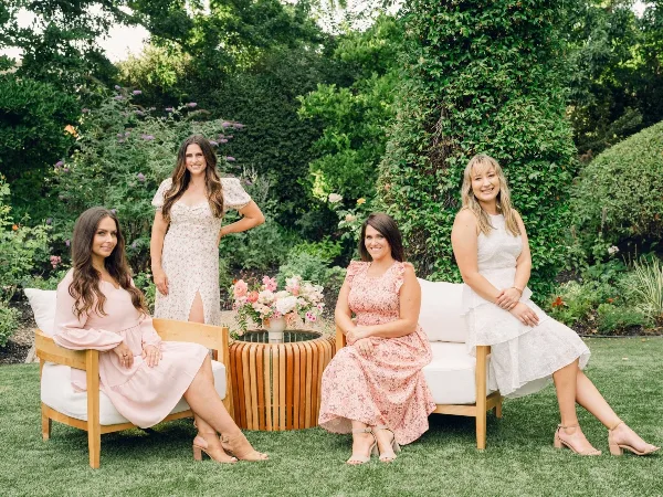 Four women in pastel dresses posing on a white outdoor sofa in a garden setting.