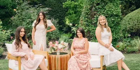 Four women in pastel dresses posing on a white outdoor sofa in a garden setting.