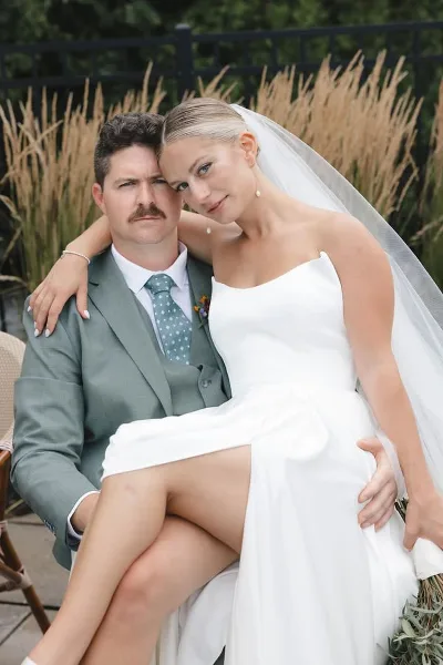 A bride with a sleek updo and natural makeup sitting on a groom's lap outdoors.