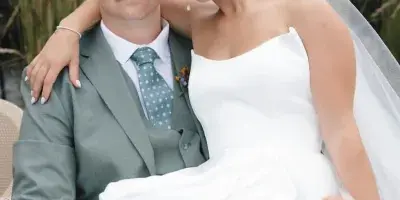 A bride with a sleek updo and natural makeup sitting on a groom's lap outdoors.