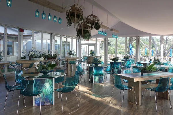 An indoor event space decorated with teal chairs, glass tables, and hanging floral arrangements.