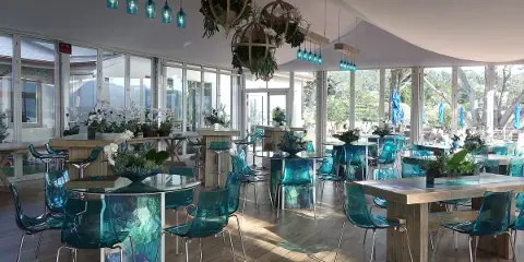 An indoor event space decorated with teal chairs, glass tables, and hanging floral arrangements.