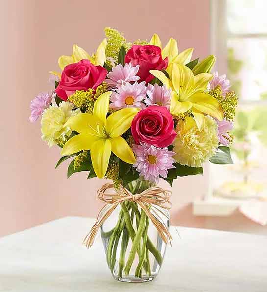 A colorful floral arrangement with red roses, yellow lilies, and pink daisies in a clear glass vase tied with twine.