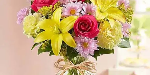 A colorful floral arrangement with red roses, yellow lilies, and pink daisies in a clear glass vase tied with twine.