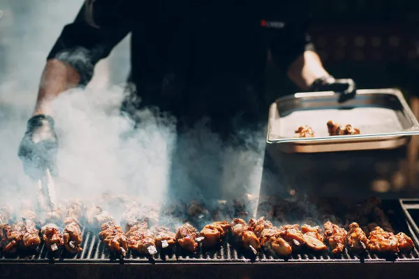 A caterer in black gloves grilling skewered meat on a large outdoor grill with smoke rising.