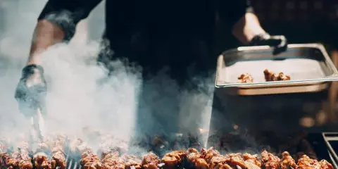 A caterer in black gloves grilling skewered meat on a large outdoor grill with smoke rising.