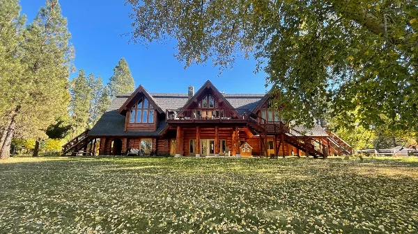 Wide exterior view of a large rustic log cabin lodge with multiple gables, balconies, and stairs set on a grassy lawn surrounded by trees under a blue sky.