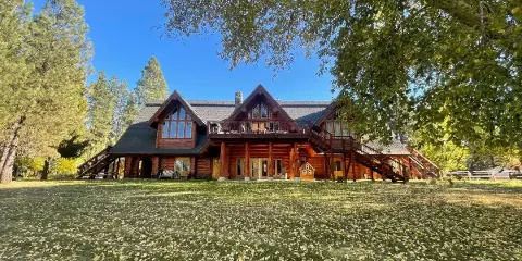 Wide exterior view of a large rustic log cabin lodge with multiple gables, balconies, and stairs set on a grassy lawn surrounded by trees under a blue sky.