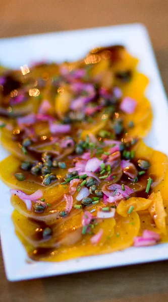 A close-up of a plated appetizer featuring sliced yellow squash topped with capers, red onions, and a dark glaze.