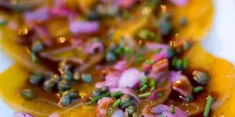 A close-up of a plated appetizer featuring sliced yellow squash topped with capers, red onions, and a dark glaze.