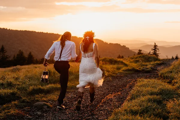 A bride and partner walking hand-in-hand on a mountain trail at sunset, with the bride holding a lantern and bouquet.