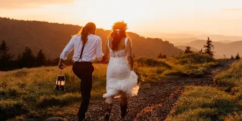A bride and partner walking hand-in-hand on a mountain trail at sunset, with the bride holding a lantern and bouquet.
