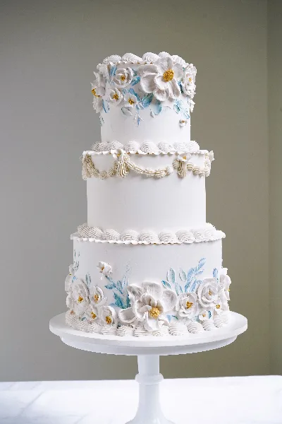 A three-tier white wedding cake decorated with hand-painted blue and white flowers and piped buttercream borders, displayed on a white pedestal stand.
