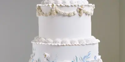 A three-tier white wedding cake decorated with hand-painted blue and white flowers and piped buttercream borders, displayed on a white pedestal stand.