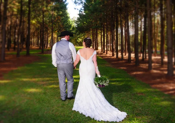 A bride and groom walking hand-in-hand through a forest of tall pine trees on a grassy path.
