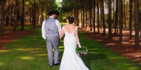 A bride and groom walking hand-in-hand through a forest of tall pine trees on a grassy path.