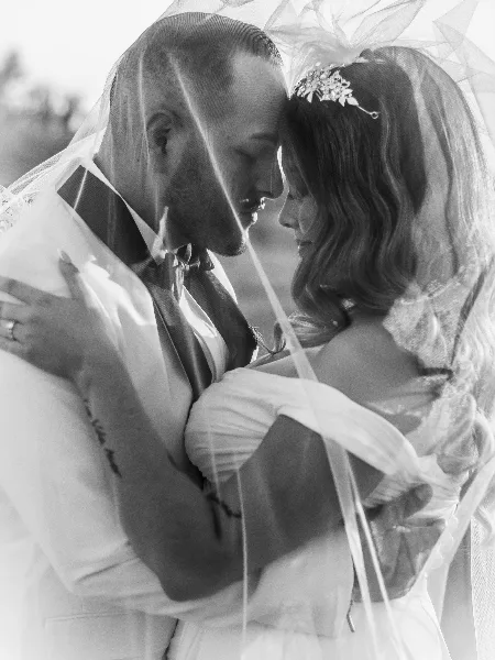 A black-and-white photograph of a bride and groom embracing, with the bride's veil draped over both of them.