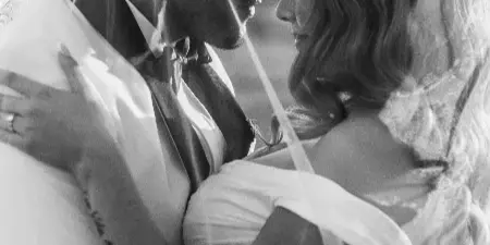 A black-and-white photograph of a bride and groom embracing, with the bride's veil draped over both of them.