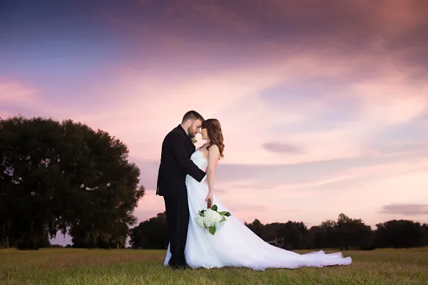 A bride and groom standing in a grassy field at sunset, with the groom kissing the bride's forehead.
