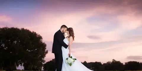 A bride and groom standing in a grassy field at sunset, with the groom kissing the bride's forehead.
