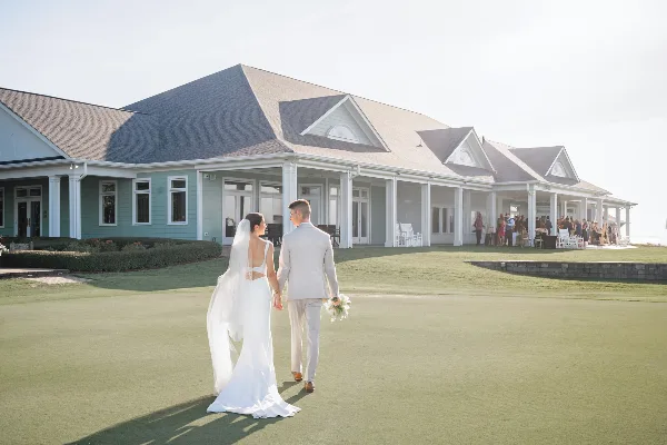 A bride and groom walking hand-in-hand across a green lawn toward a coastal wedding venue building.