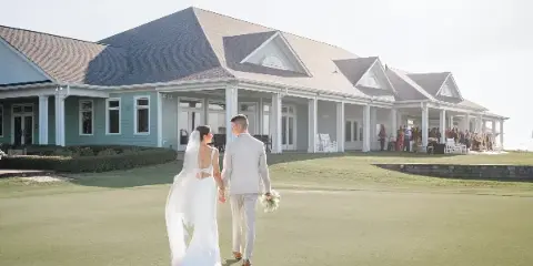 A bride and groom walking hand-in-hand across a green lawn toward a coastal wedding venue building.