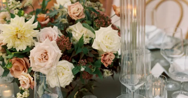 Close-up of a lush wedding floral centerpiece featuring cream and blush roses, dahlias, and greenery on a table with glassware and candles.