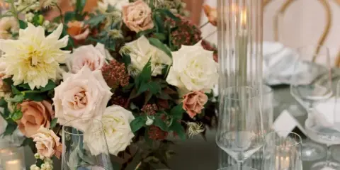 Close-up of a lush wedding floral centerpiece featuring cream and blush roses, dahlias, and greenery on a table with glassware and candles.