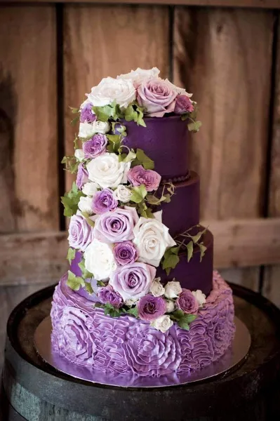 A four-tier purple and lavender wedding cake decorated with fresh white and purple roses and greenery, displayed on a wooden barrel.