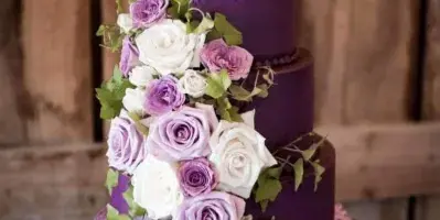 A four-tier purple and lavender wedding cake decorated with fresh white and purple roses and greenery, displayed on a wooden barrel.