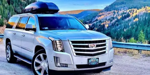 A white luxury SUV with a Blue Sky Limo plate frame parked on a mountain road with forested hills in the background.