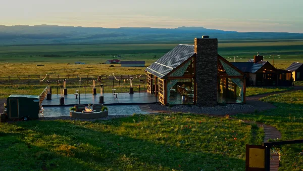 A wide shot of a modern glass and wood pavilion with a stone chimney, situated on a grassy ranch property with a view of distant mountains at sunset.