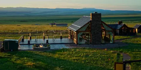 A wide shot of a modern glass and wood pavilion with a stone chimney, situated on a grassy ranch property with a view of distant mountains at sunset.