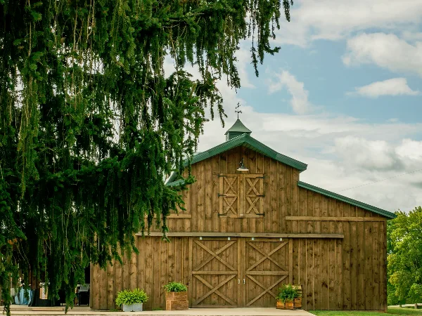 Exterior view of a large wooden barn with a green metal roof and a cupola, set against a blue sky with white clouds.