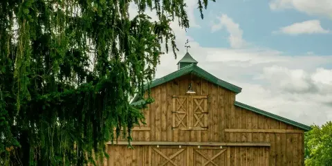 Exterior view of a large wooden barn with a green metal roof and a cupola, set against a blue sky with white clouds.