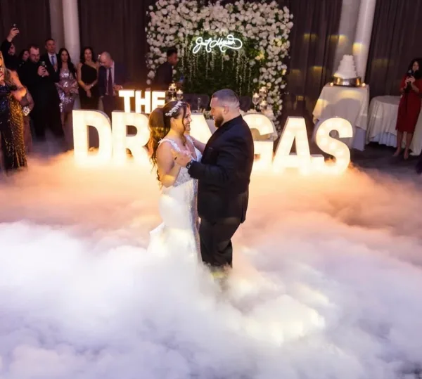 A bride and groom dancing on a fog-covered dance floor at a wedding reception with illuminated marquee letters in the background.