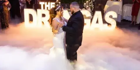 A bride and groom dancing on a fog-covered dance floor at a wedding reception with illuminated marquee letters in the background.