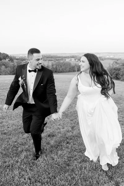 A black and white photograph of a bride and groom holding hands and walking through a grassy field.