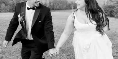 A black and white photograph of a bride and groom holding hands and walking through a grassy field.