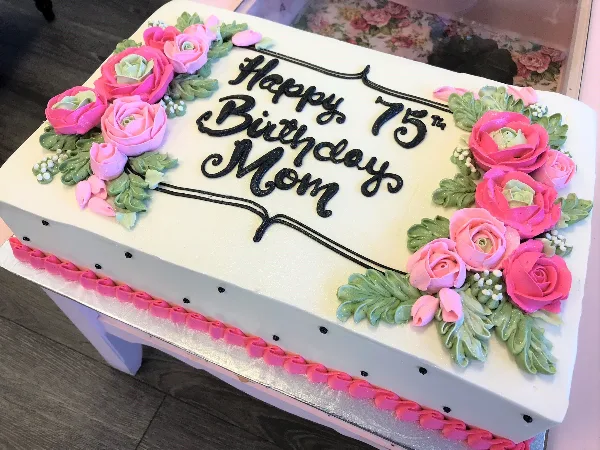 A white sheet cake decorated with pink and green piped roses and black lettering reading Happy 75th Birthday Mom.