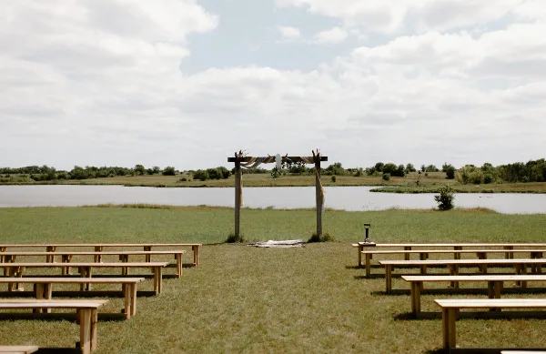 An outdoor wedding ceremony setup with a wooden arch and rows of benches on a grassy field next to a large pond.