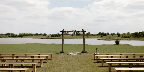 An outdoor wedding ceremony setup with a wooden arch and rows of benches on a grassy field next to a large pond.