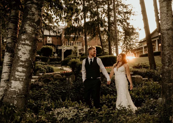 A bride and groom walking hand-in-hand through the gardens of a historic estate venue at sunset.