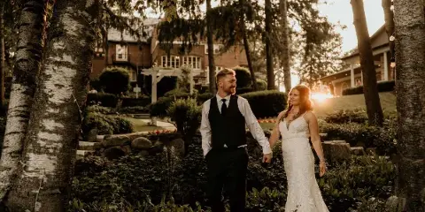 A bride and groom walking hand-in-hand through the gardens of a historic estate venue at sunset.