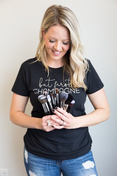 A smiling blonde woman wearing a black t-shirt holds a set of professional makeup brushes.