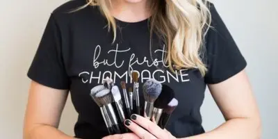 A smiling blonde woman wearing a black t-shirt holds a set of professional makeup brushes.