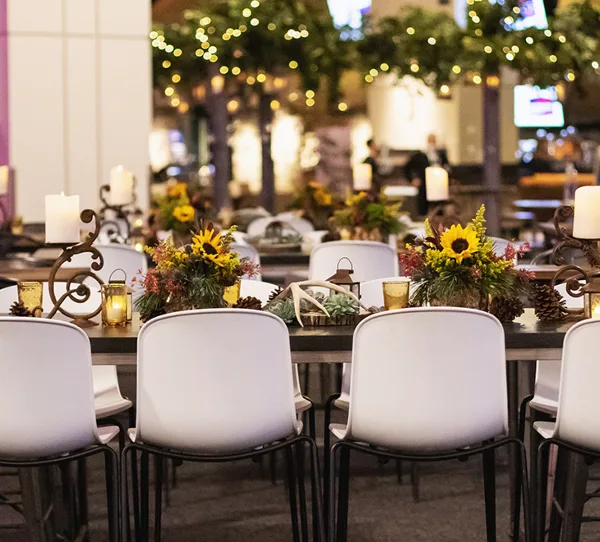 A close-up view of a decorated event table featuring white chairs, sunflower centerpieces, and candlelight.